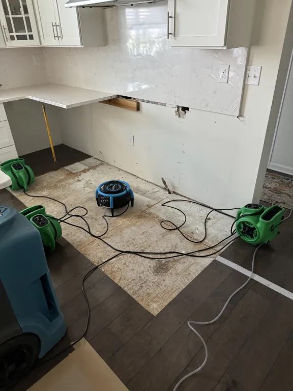 Kitchen with a section of flooring removed to expose the subfloor, with air movers and power cords on the floor.