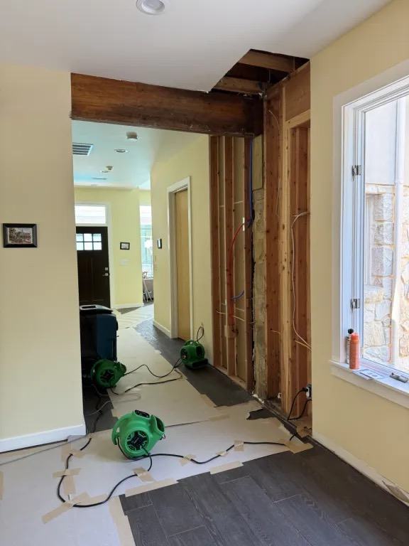 Hallway with one wall opened to the studs, an opening in the ceiling, paper floor protection, and green air movers.